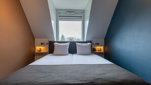 Un dormitorio con tragaluz, cama con ropa de cama blanca, dos lámparas de noche y vista a árboles por la ventana.