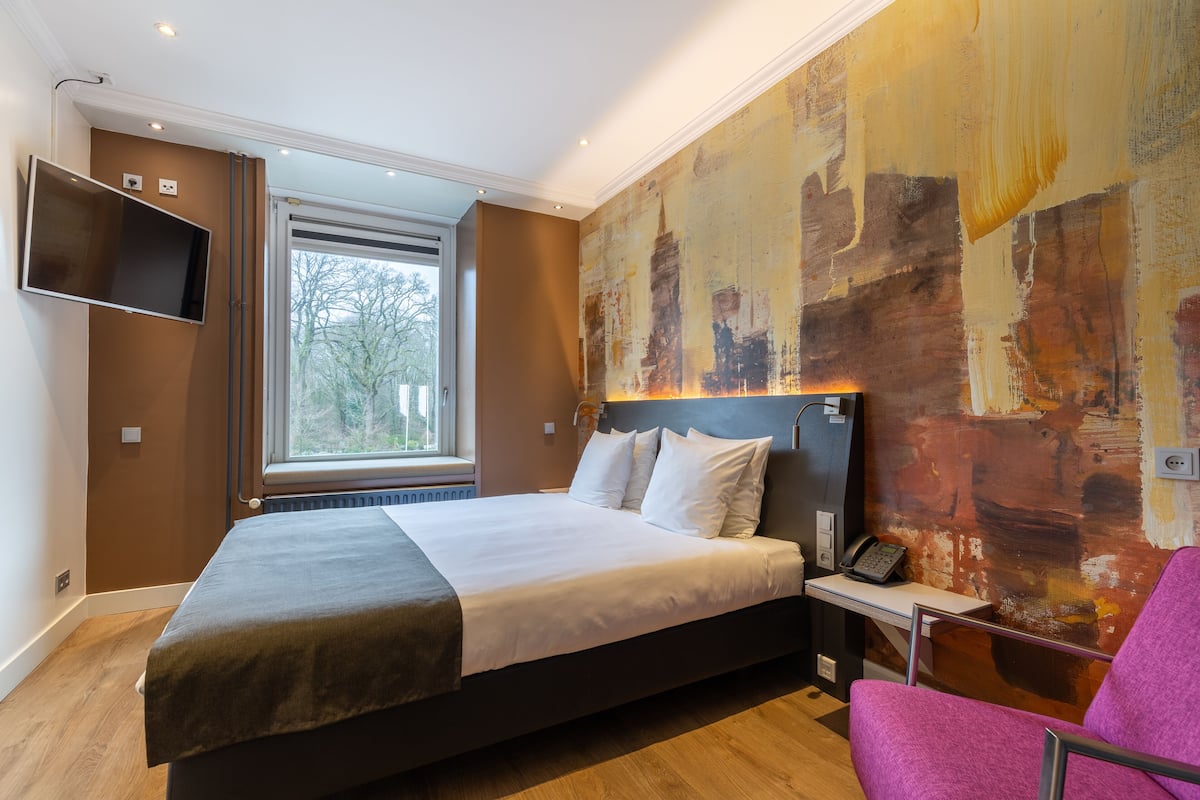 Una habitación de hotel moderna con una cama grande, un televisor de pantalla plana en la pared, una ventana con vistas a árboles y un cuadro abstracto de gran tamaño en la pared.