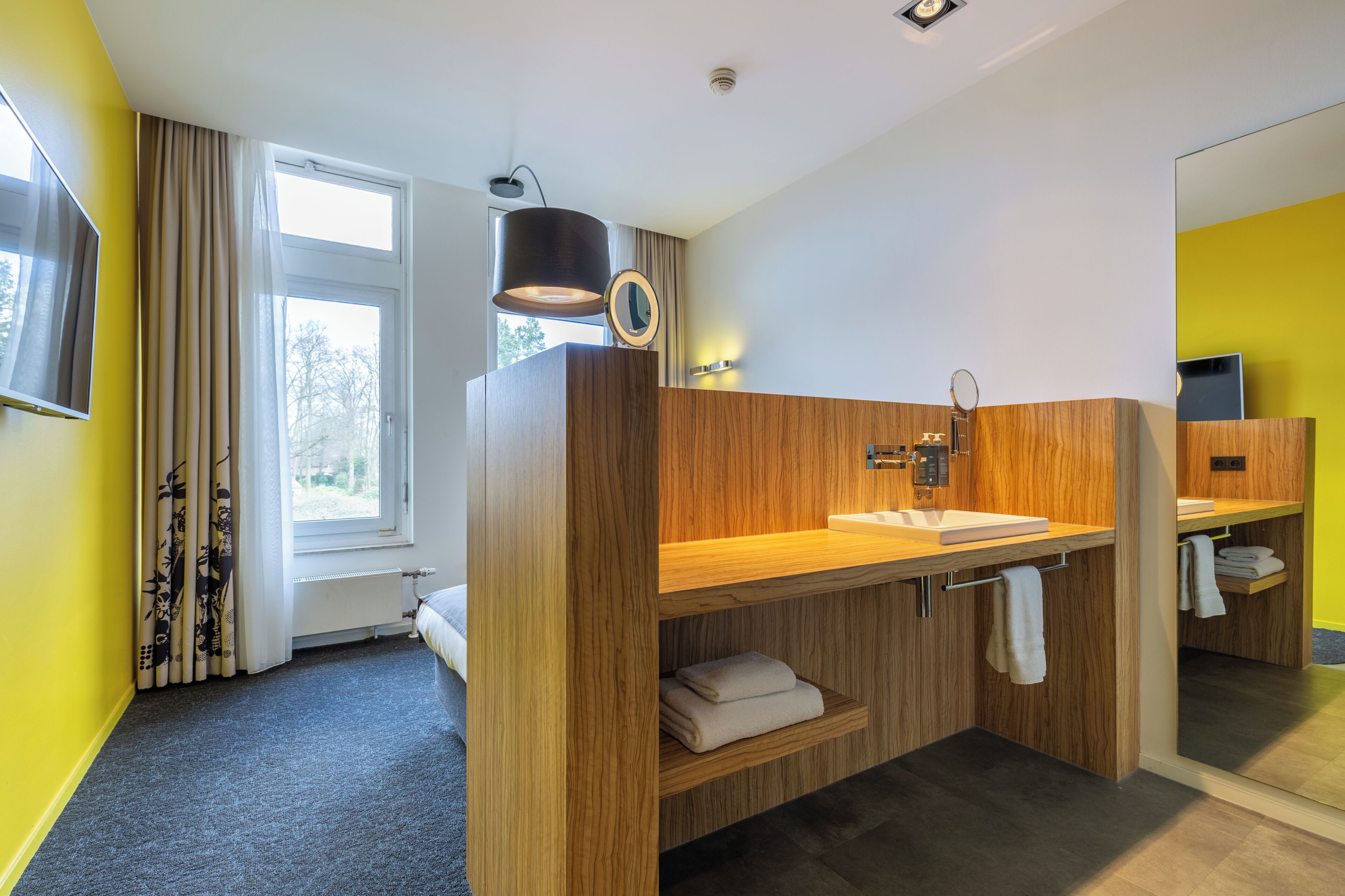 A modern bathroom with wooden cabinetry, a large mirror, and a window with curtains.