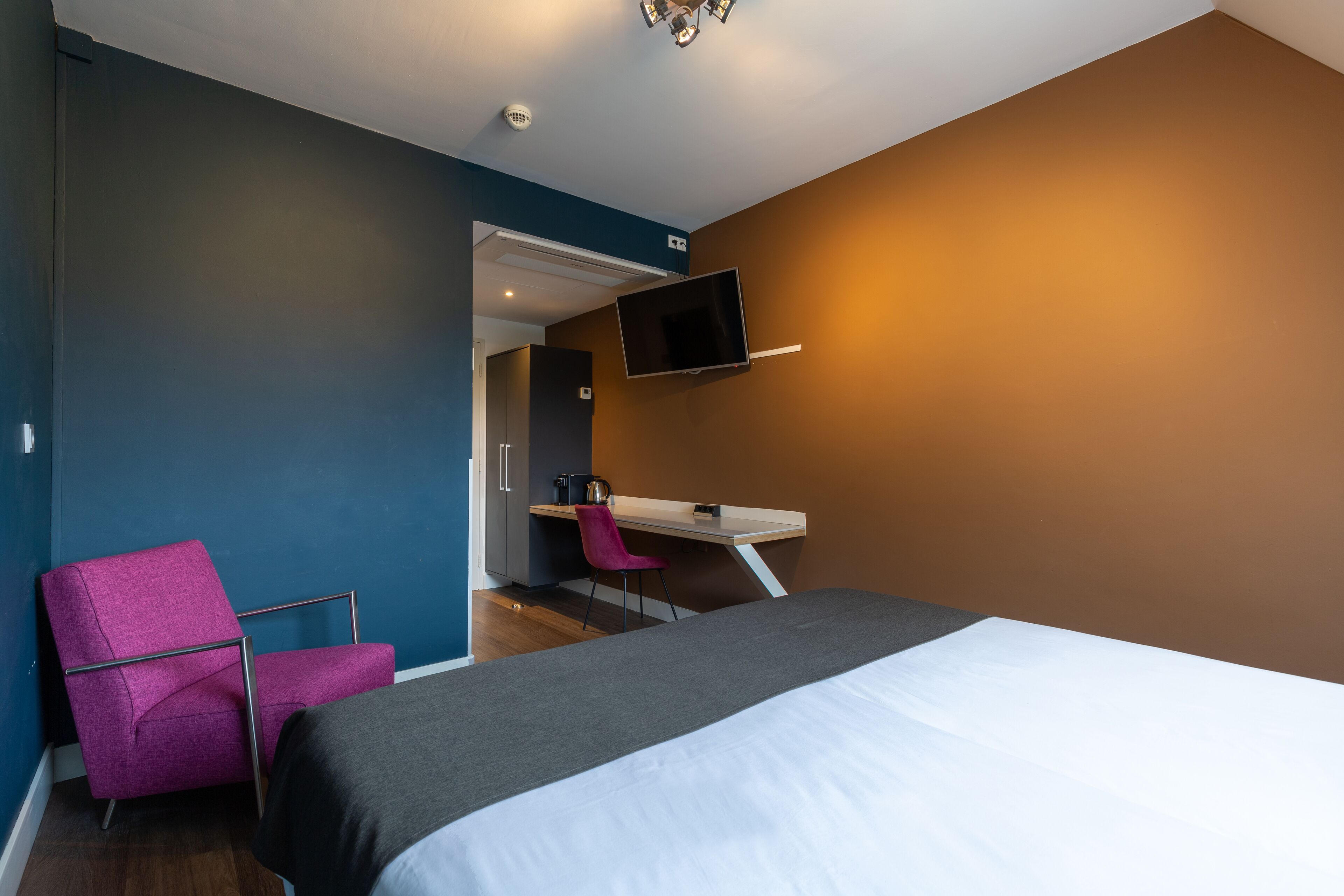 A modern hotel room with a bed, a desk, a chair, and a TV mounted on the wall.
