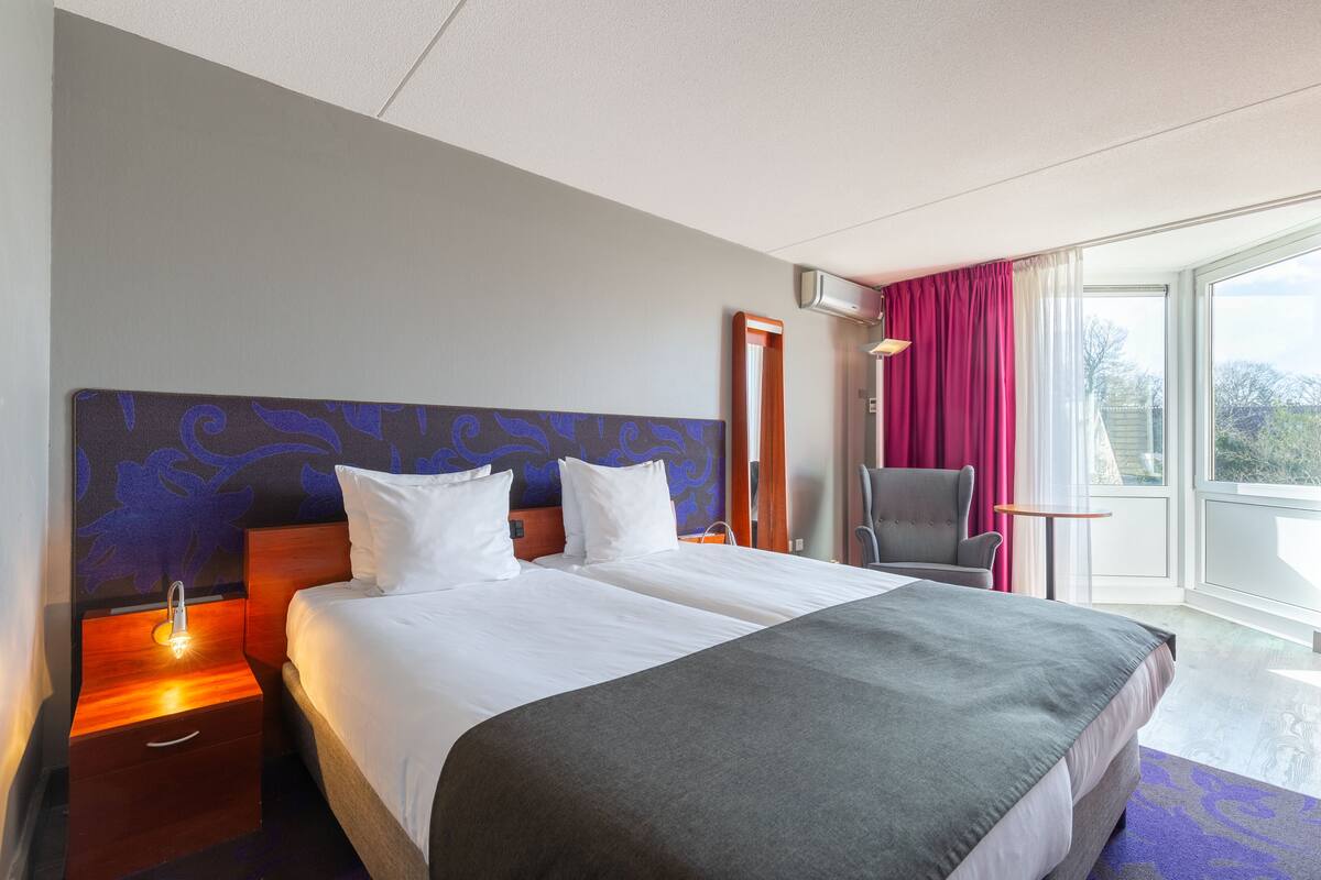 Una habitación de hotel con una cama grande, una mesita de noche con lámpara, una silla y una ventana con cortinas.