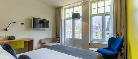 A hotel room with a bed, a desk, a chair, a TV, and large windows.
