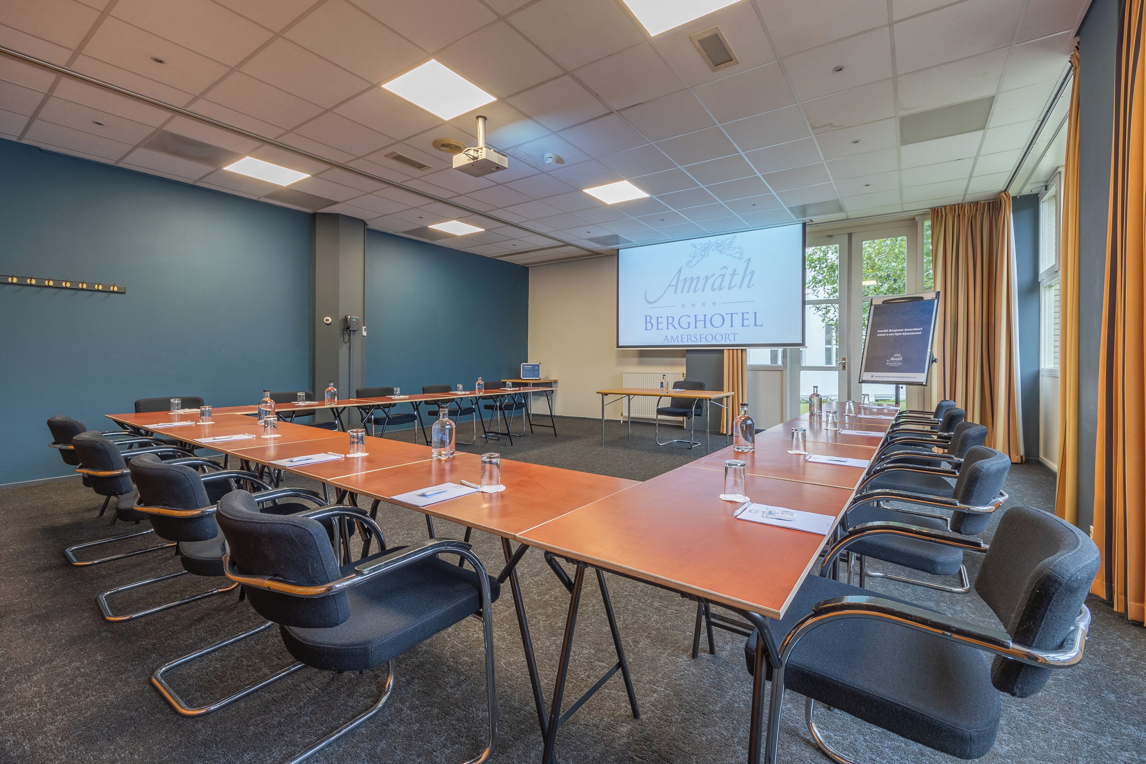 Een vergaderruimte met een lange tafel, stoelen, een projectiescherm en een presentatie over Amrath Berghotel Amersfoort.