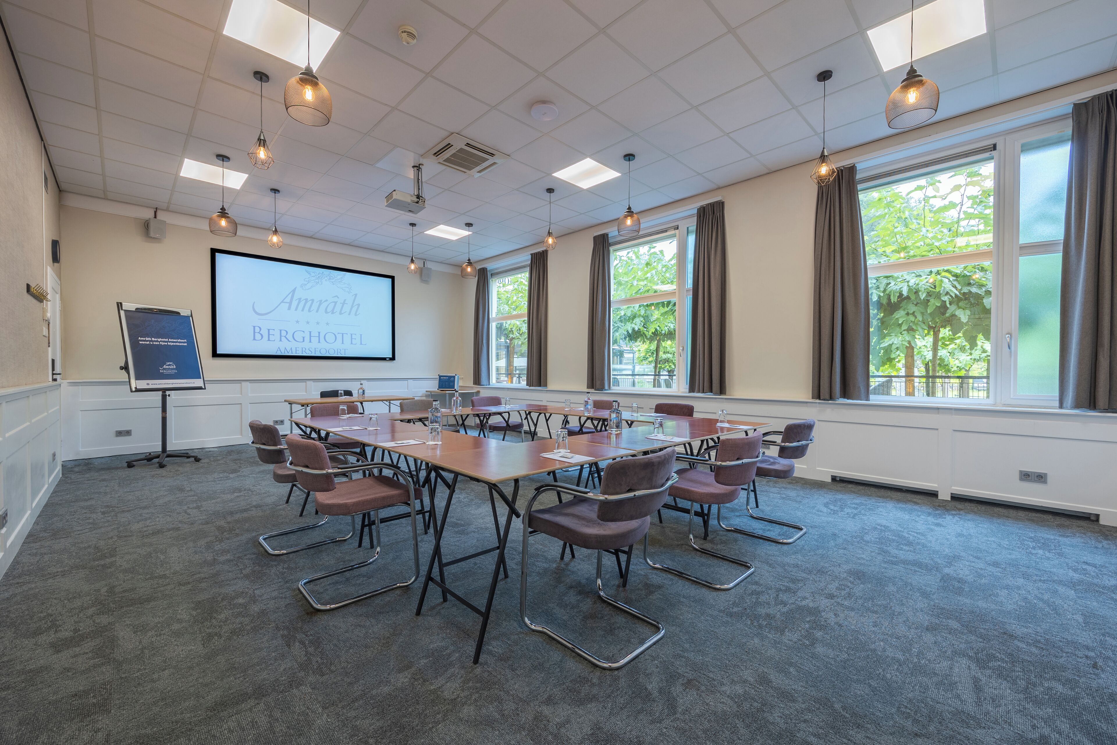 A conference room with a large table, chairs, a projector screen, and windows with curtains.