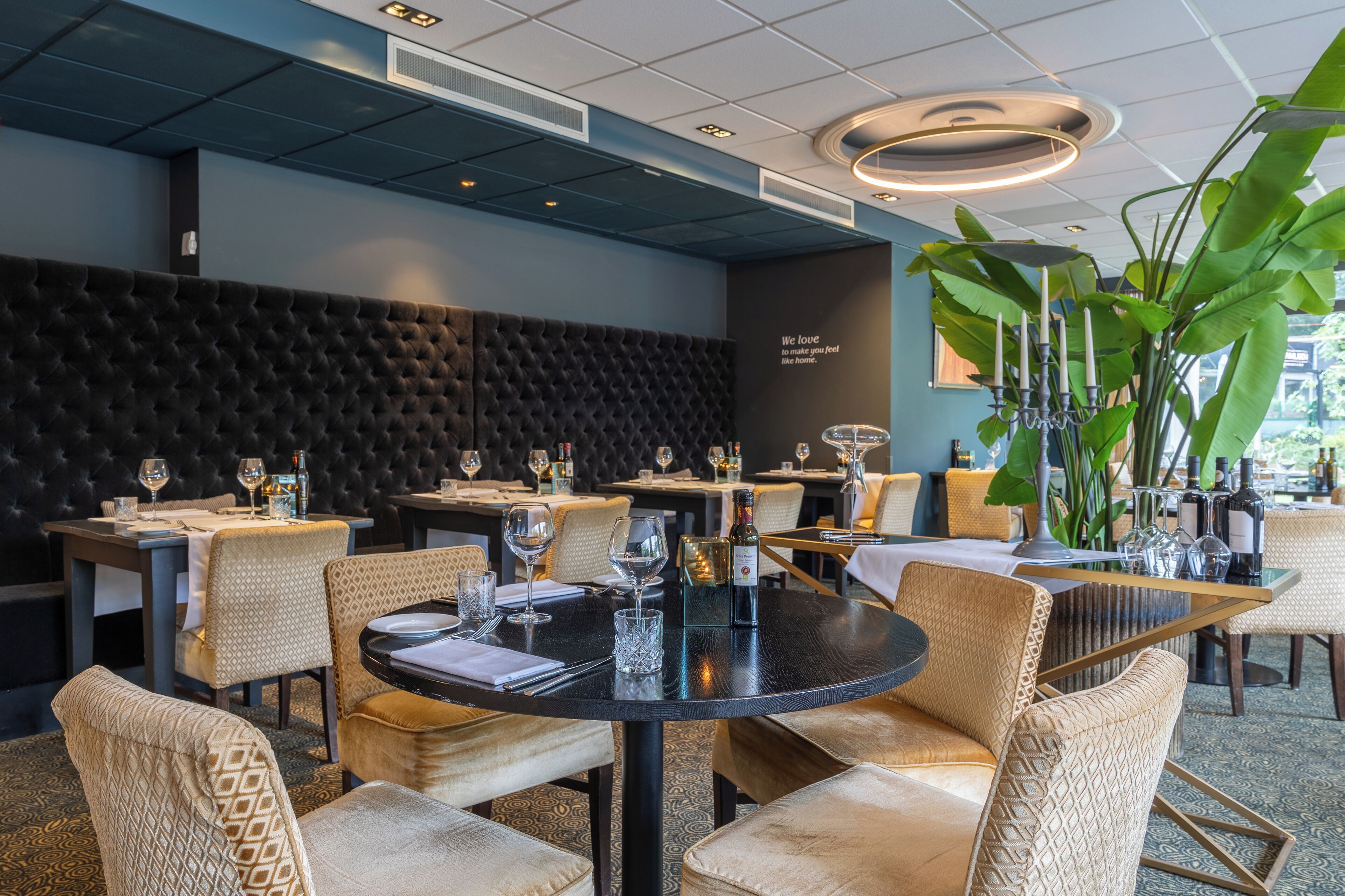 A restaurant interior with round tables, cushioned chairs, a black upholstered wall, and a large green plant.