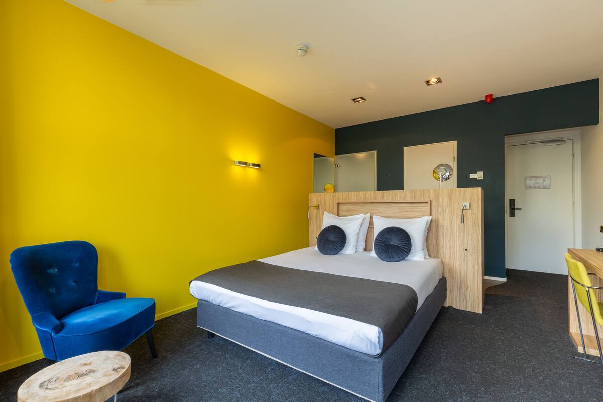 Una habitación de hotel con una cama, una silla azul, un escritorio de madera y un espejo.