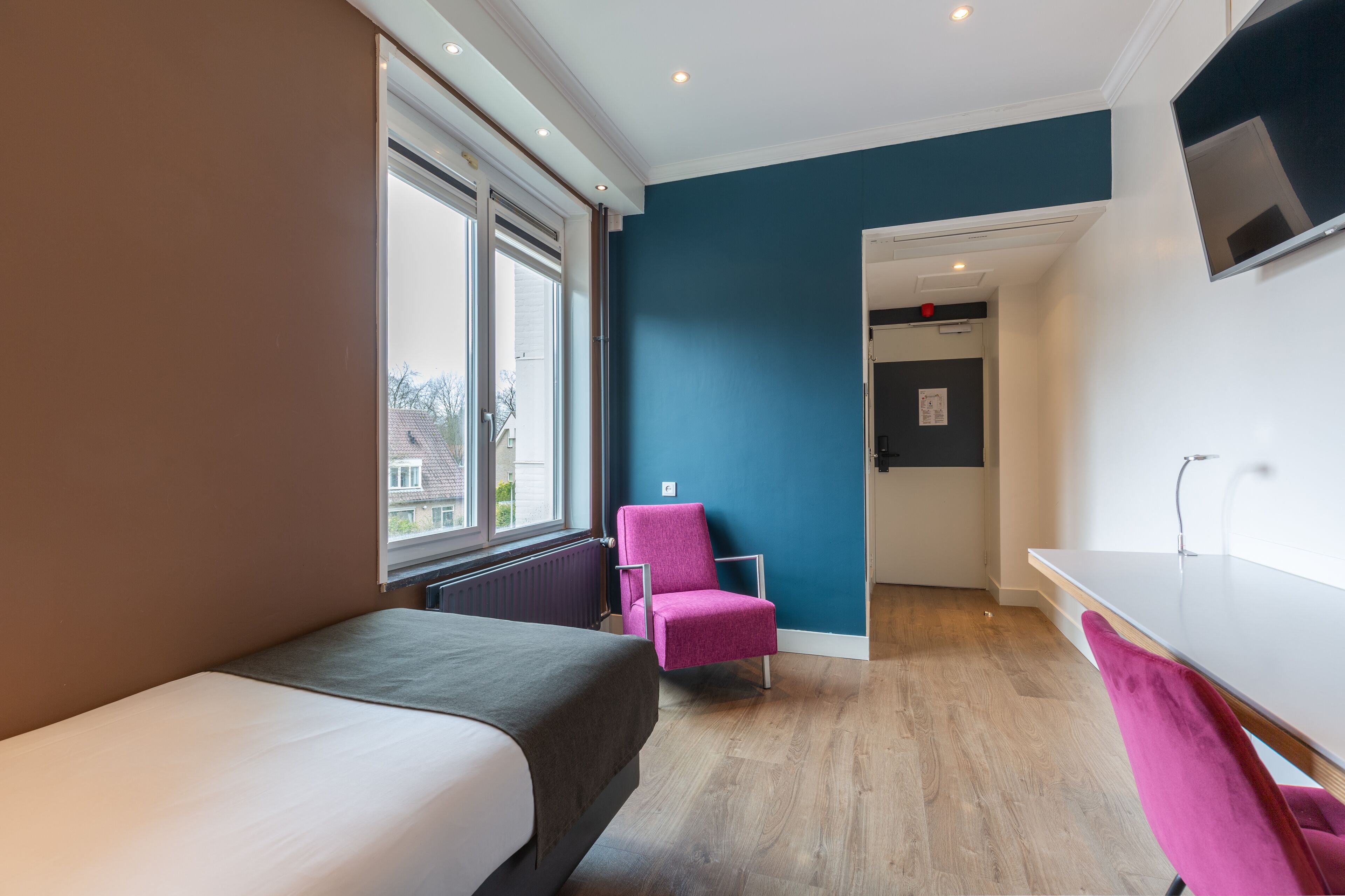 Een moderne hotelkamer met een bed, een roze stoel, een bureau en een aan de muur bevestigde televisie.