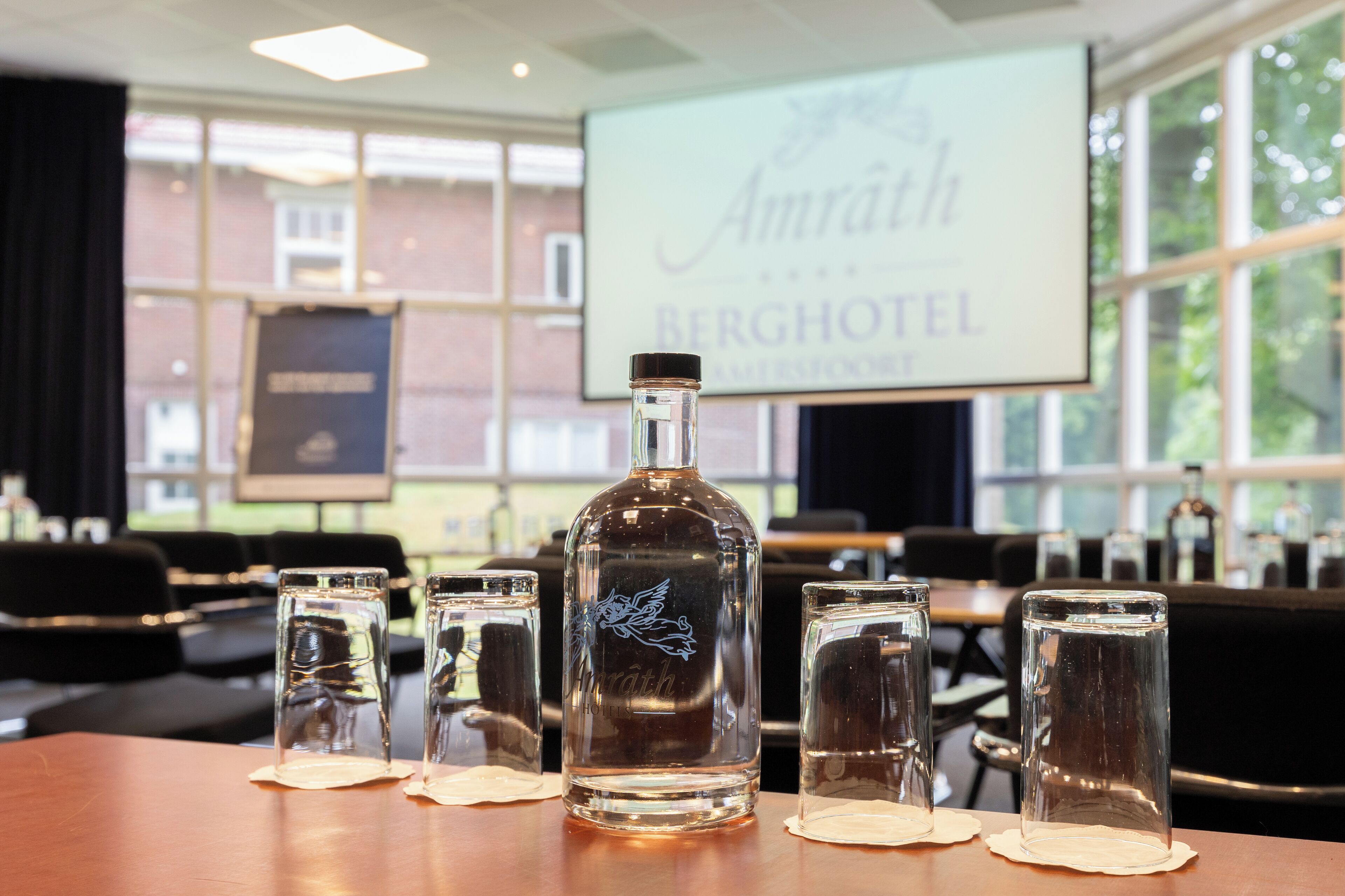 A bottle of Amrath liquor and four glasses on a table in a hotel room with a large window and a screen displaying the hotel name.