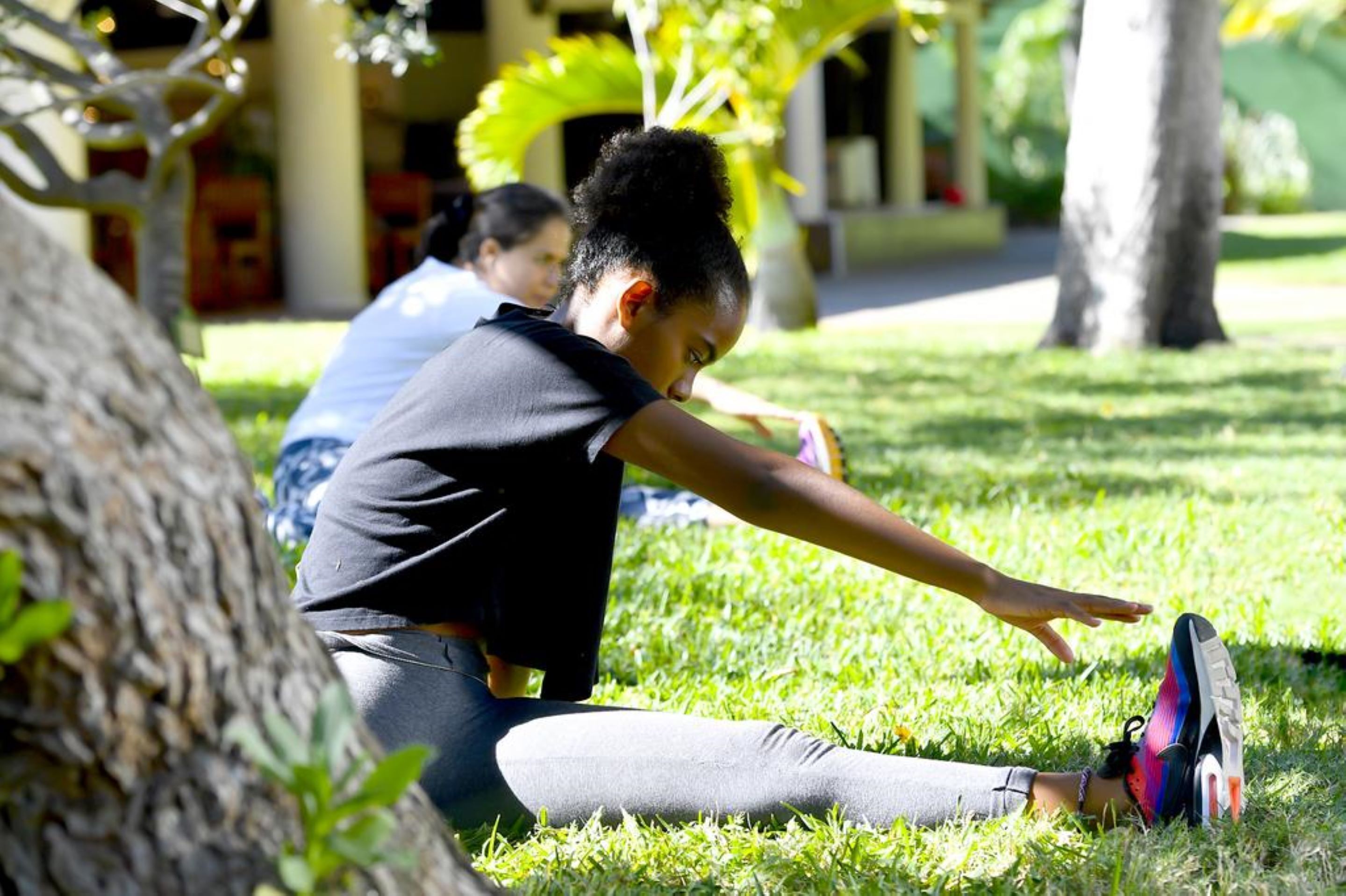 A person stretching outdoors on grass with trees and a building in the background.