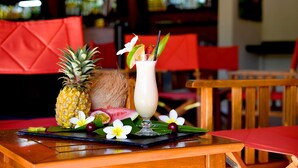 A table with a pineapple, coconut, and a drink garnished with fruit and flowers, set on a black placemat with a red tablecloth. - Le Relais de L'Hermitage (Saint-Paul)