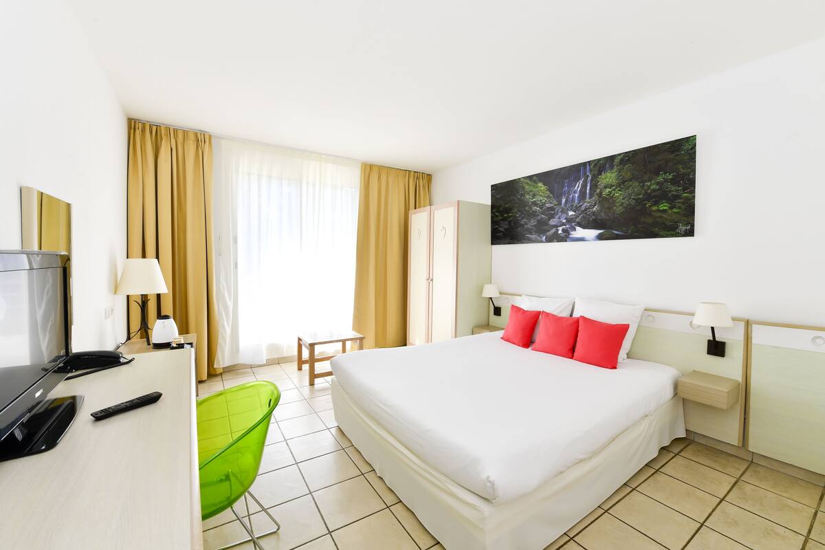 Habitación de hotel con una cama, un escritorio con computadora, una silla verde, un televisor y un cuadro de una cascada.