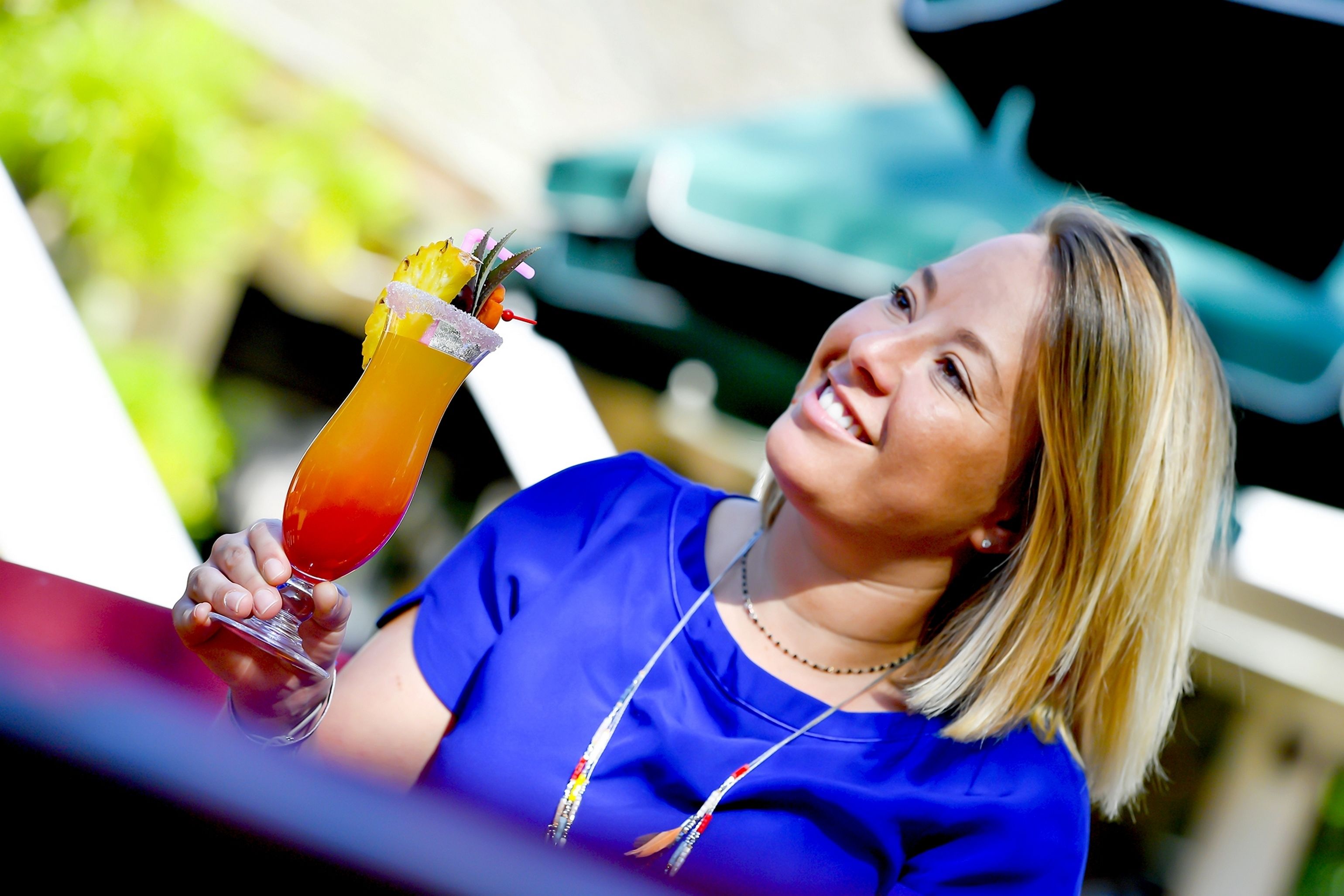 A person holding a cocktail with a garnish, seated at an outdoor table with a green umbrella in the background.
