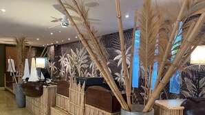 A reception area with a bamboo reception desk, a large vase with tall plants, and a wall with tropical leaf patterns. - Le Relais de L'Hermitage (Saint-Paul)