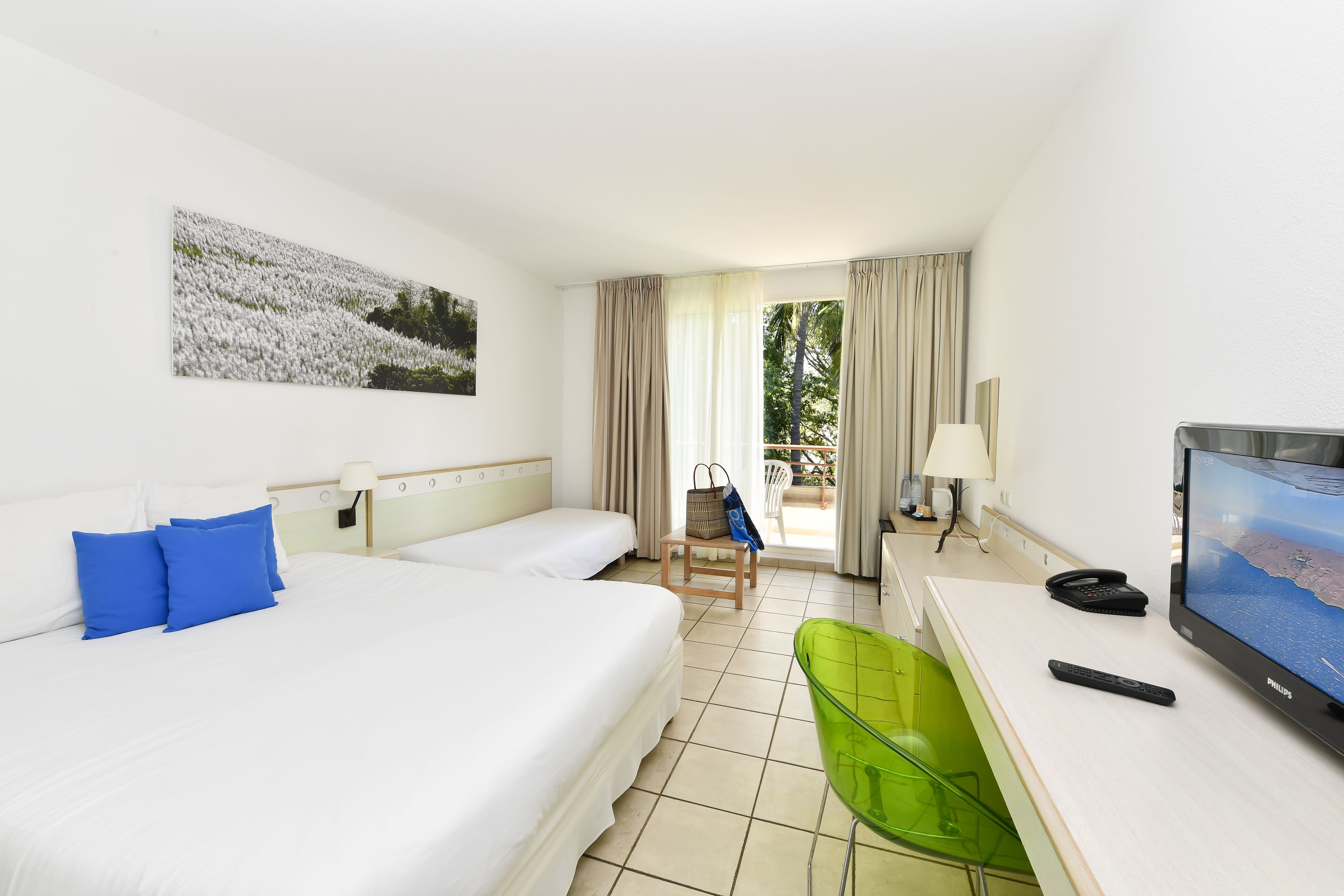 A hotel room with two beds, a desk with a computer and a TV, a green chair, and a window with curtains.