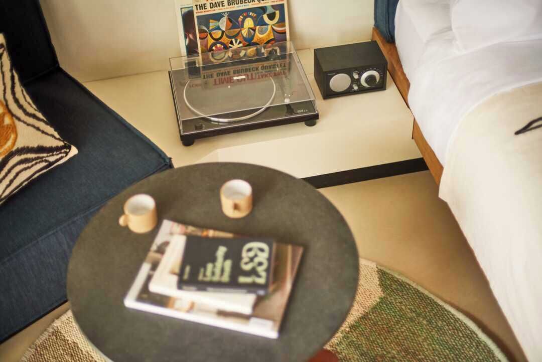 Hotellihuone, jossa on sänky, yöpöytä, jossa on radio, pieni pöytä, jossa on lehti, ja seinällä koriste-esine.