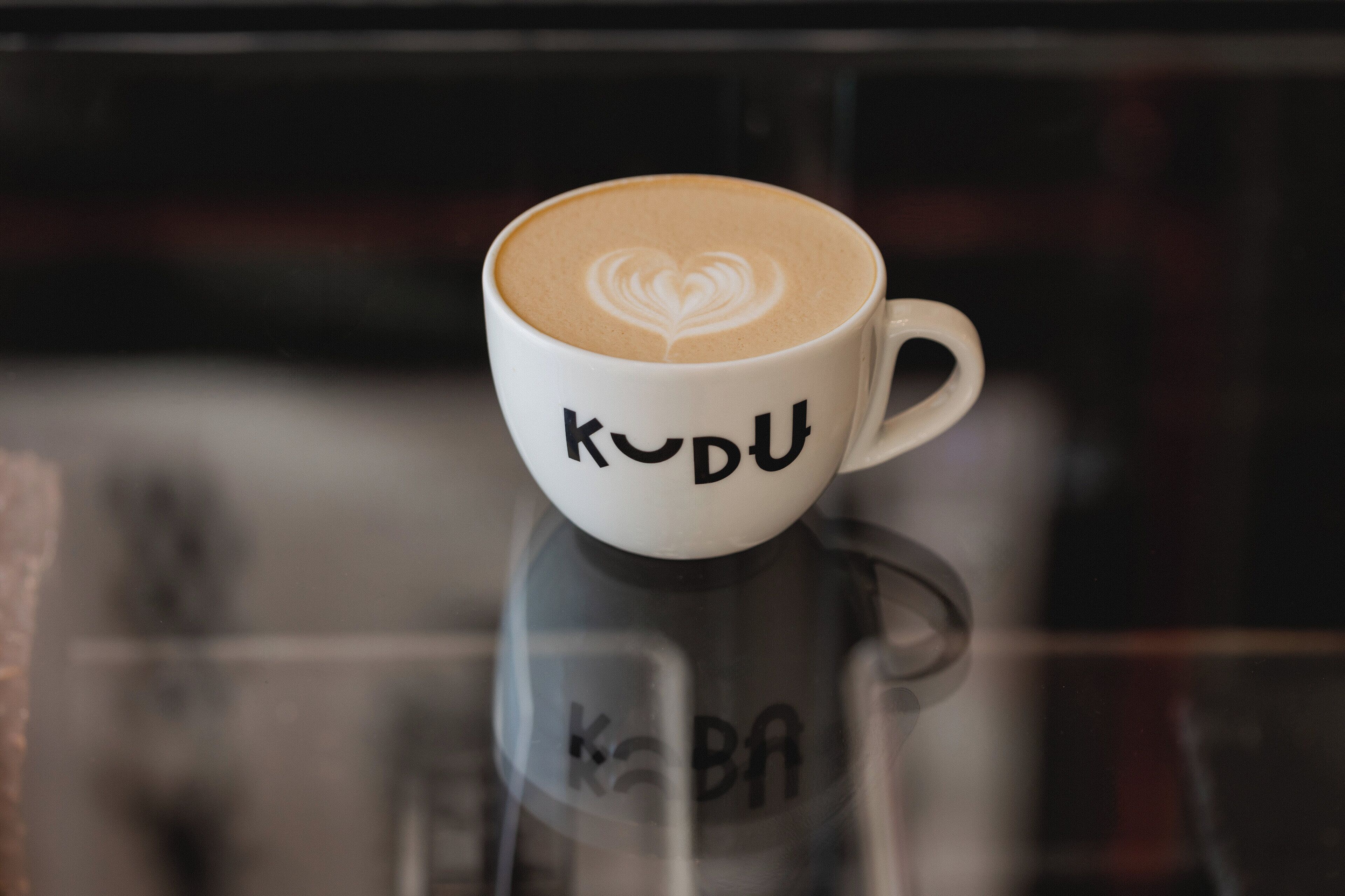 A white cup with "KUDO" and a heart design on the foam, placed on a reflective surface.