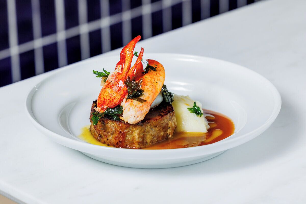 Une assiette blanche sur laquelle on trouve une queue de homard, un morceau de viande et quelques légumes verts.