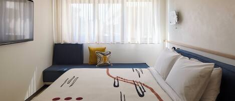 A hotel room with a bed, a window with sheer curtains, a wall-mounted TV, and a bedside table with a lamp.