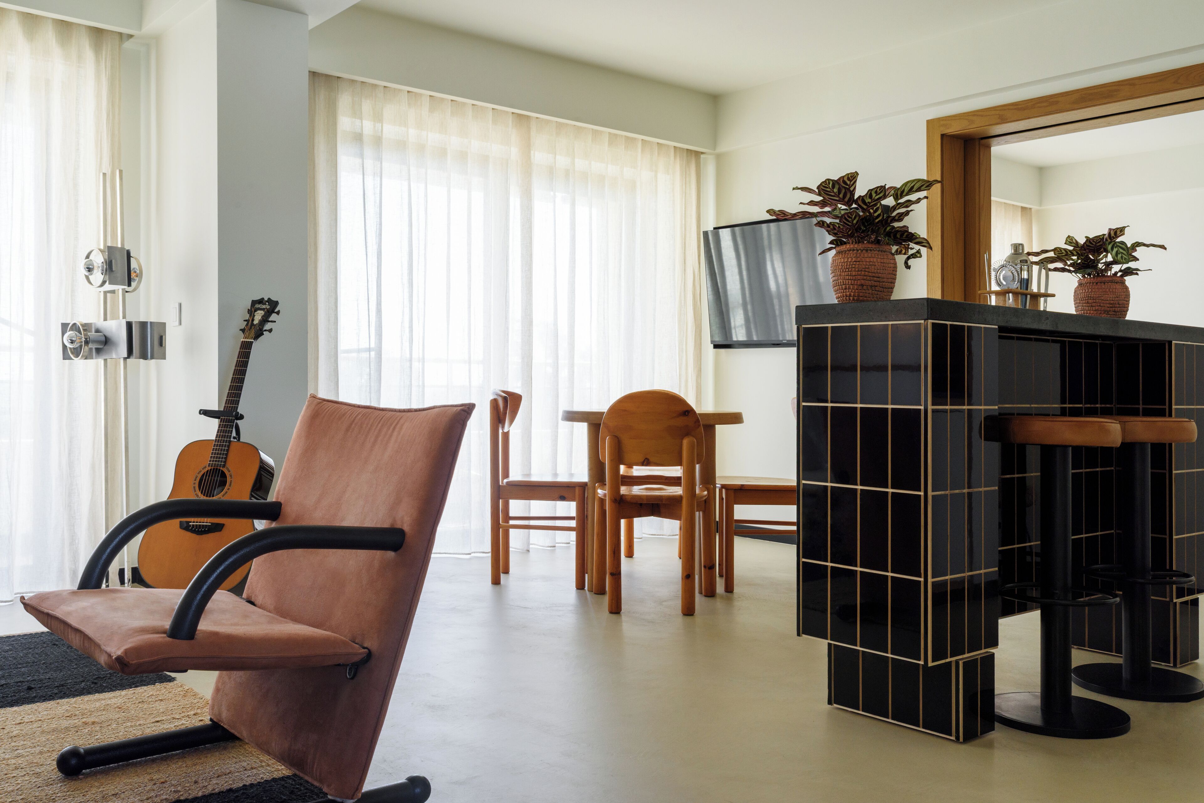 Un salón moderno con un sillón de cuero marrón, una mesa de comedor de madera con sillas, un aparador negro y dorado, y una guitarra apoyada contra la pared.