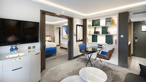 A hotel room with a flat-screen TV, a desk with a sink, a round table with chairs, a blue sofa, and a bookshelf. - Hilton London Heathrow Airport Hotel (Hounslow)