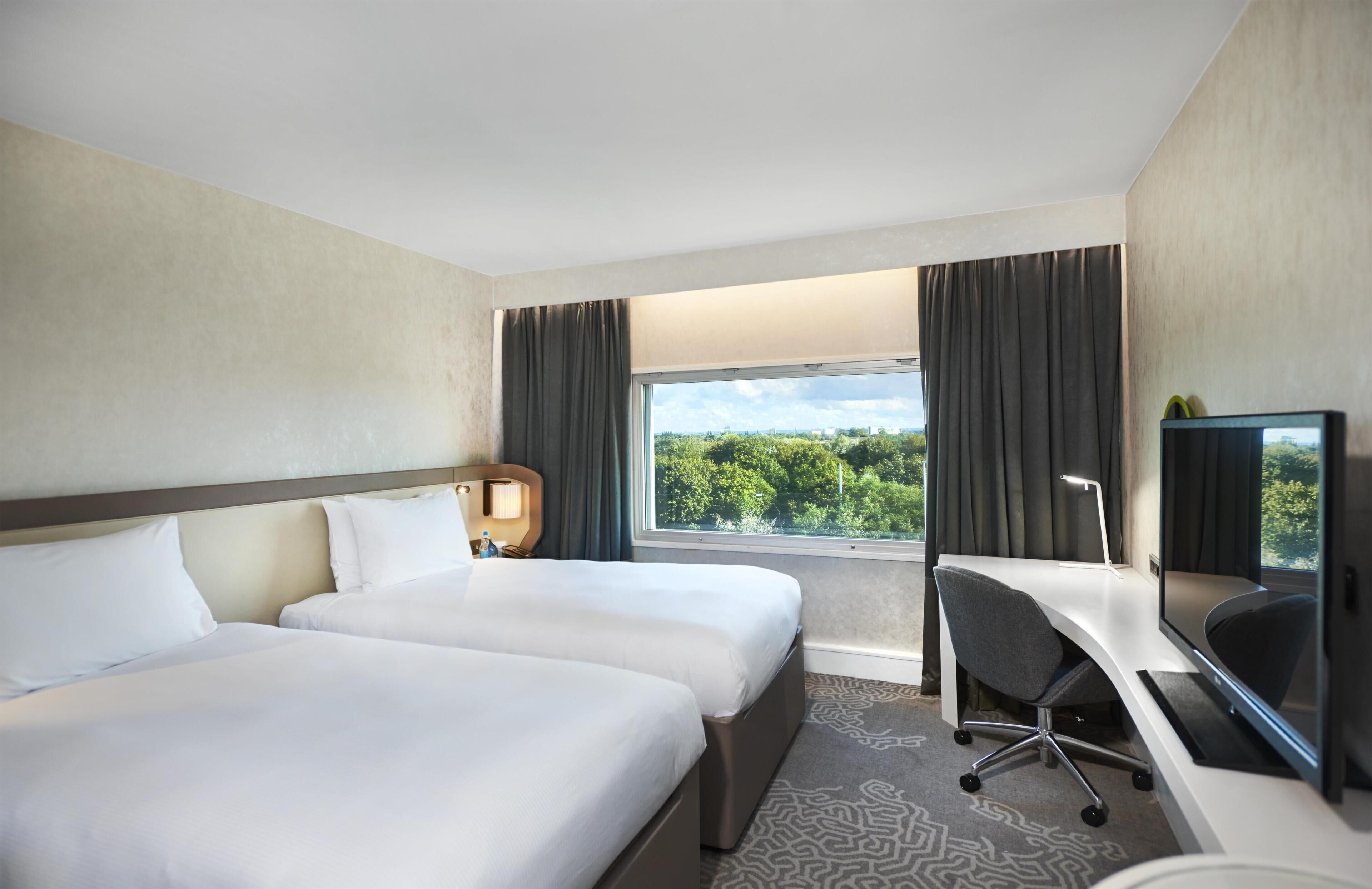 A hotel room with two beds, a desk, a chair, a TV, and a window with a view of trees.