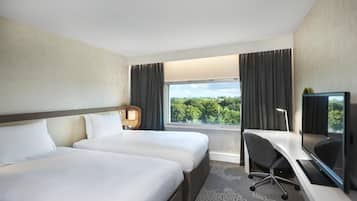 A hotel room with two beds, a desk, a chair, a TV, and a window with a view of trees.
