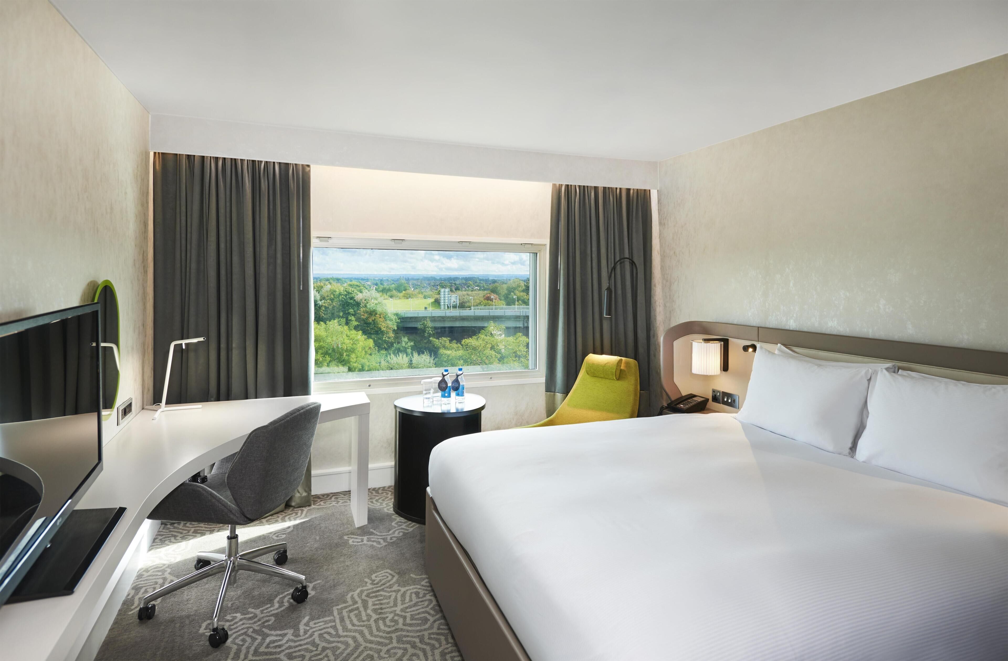 A hotel room with a bed, desk, chair, and a view of greenery through the window.