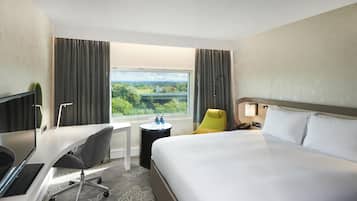 A hotel room with a bed, desk, chair, and a view of greenery through the window.