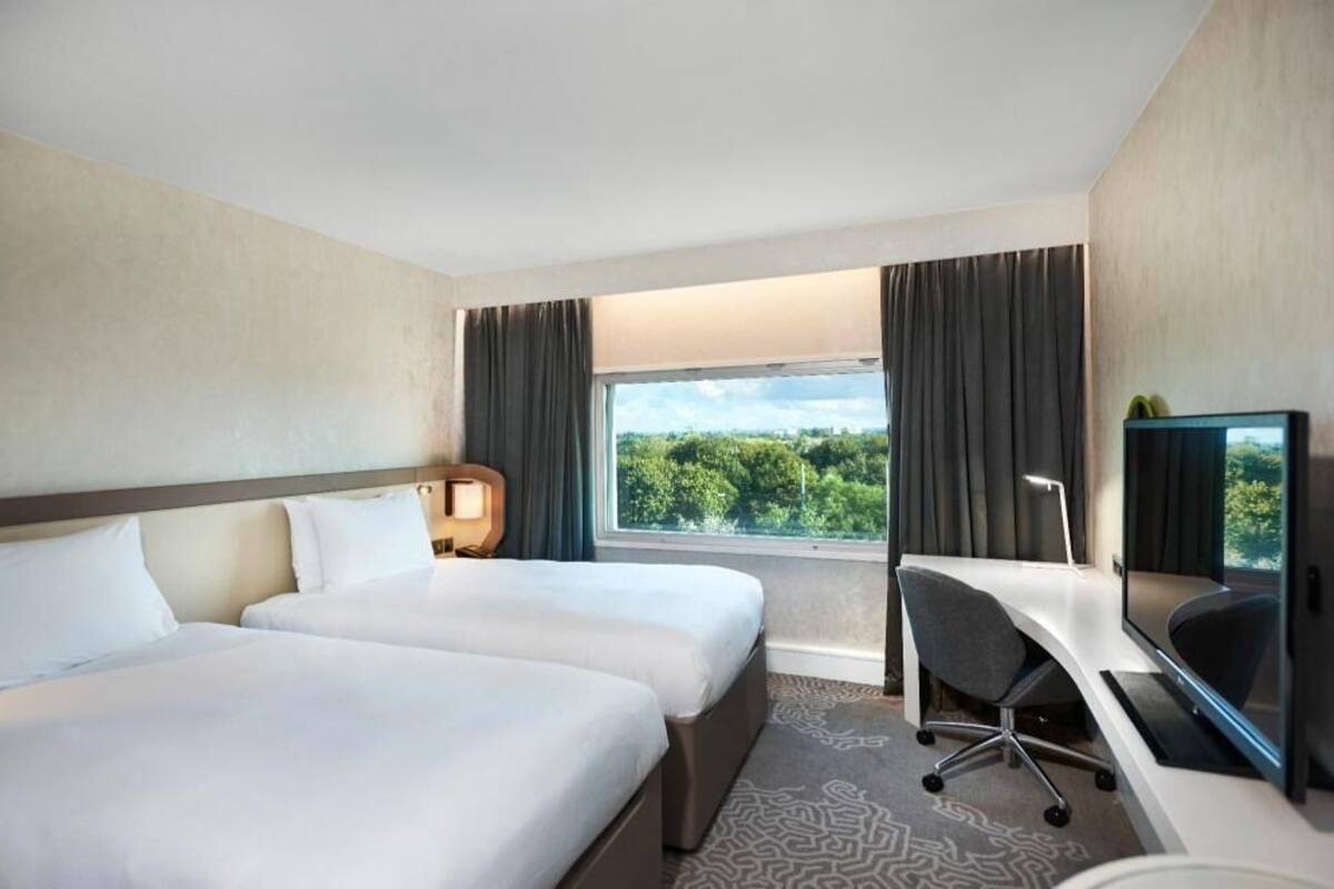Hotellihuone, jossa on kaksi sänkyä, työpöytä, tuoli, televisio ja suuri ikkuna, josta avautuu näkymä vehreään maisemaan.