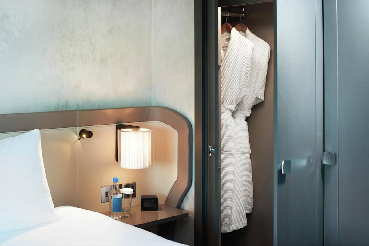 Hotellihuone, jossa on sänky, yöpöytä lampun kanssa, pullo ja kello.