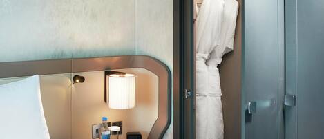 A hotel room with a bed, a bedside table with a lamp, a bottle, and a clock.