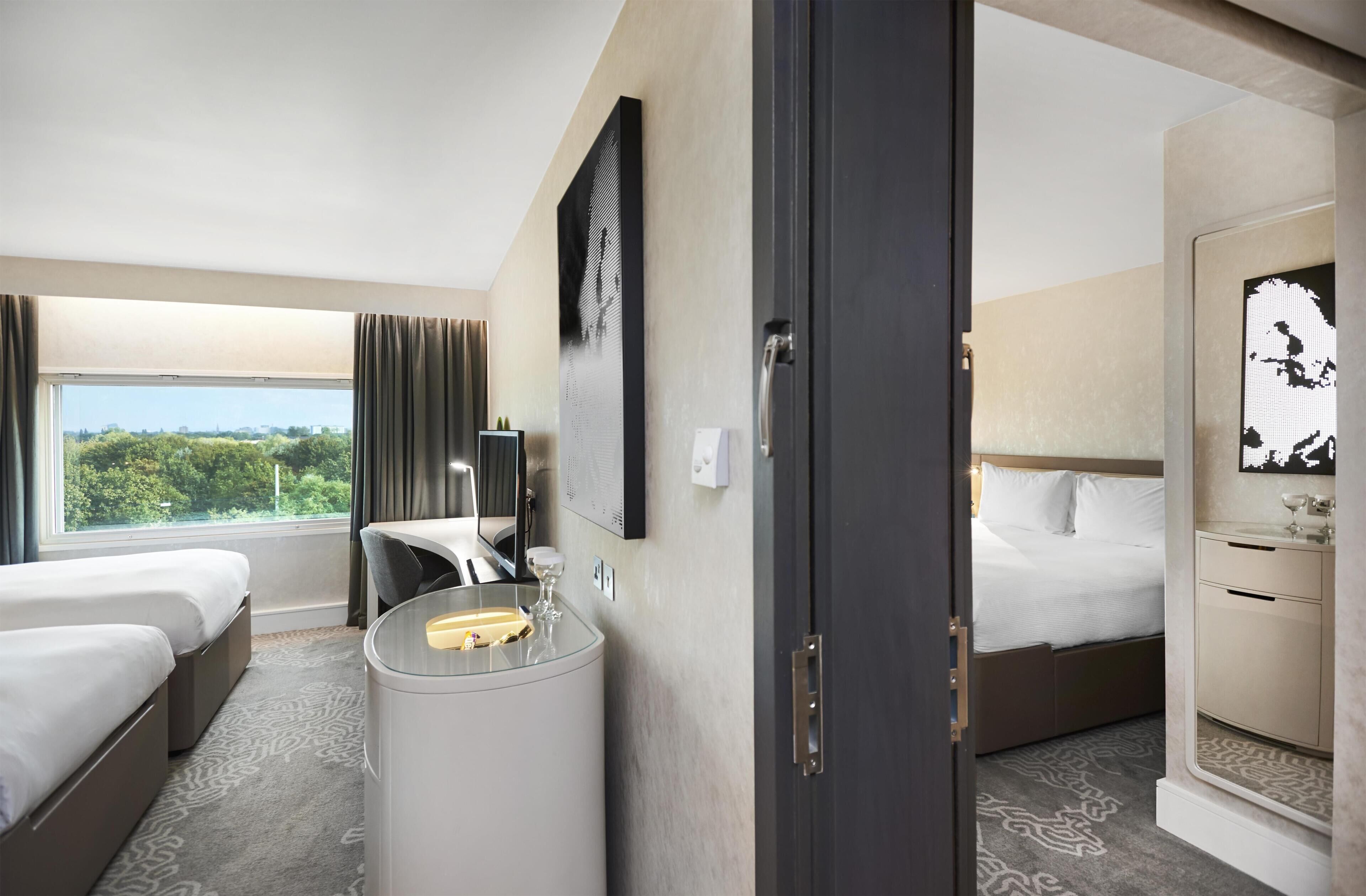 A hotel room with two beds, a minibar, a large window with curtains, a wall-mounted TV, and a view of greenery outside.