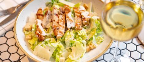 Une salade composée de poulet grillé et de croûtons, accompagnée d’un verre de vin blanc, est servie sur une table dont le plateau présente un motif hexagonal.