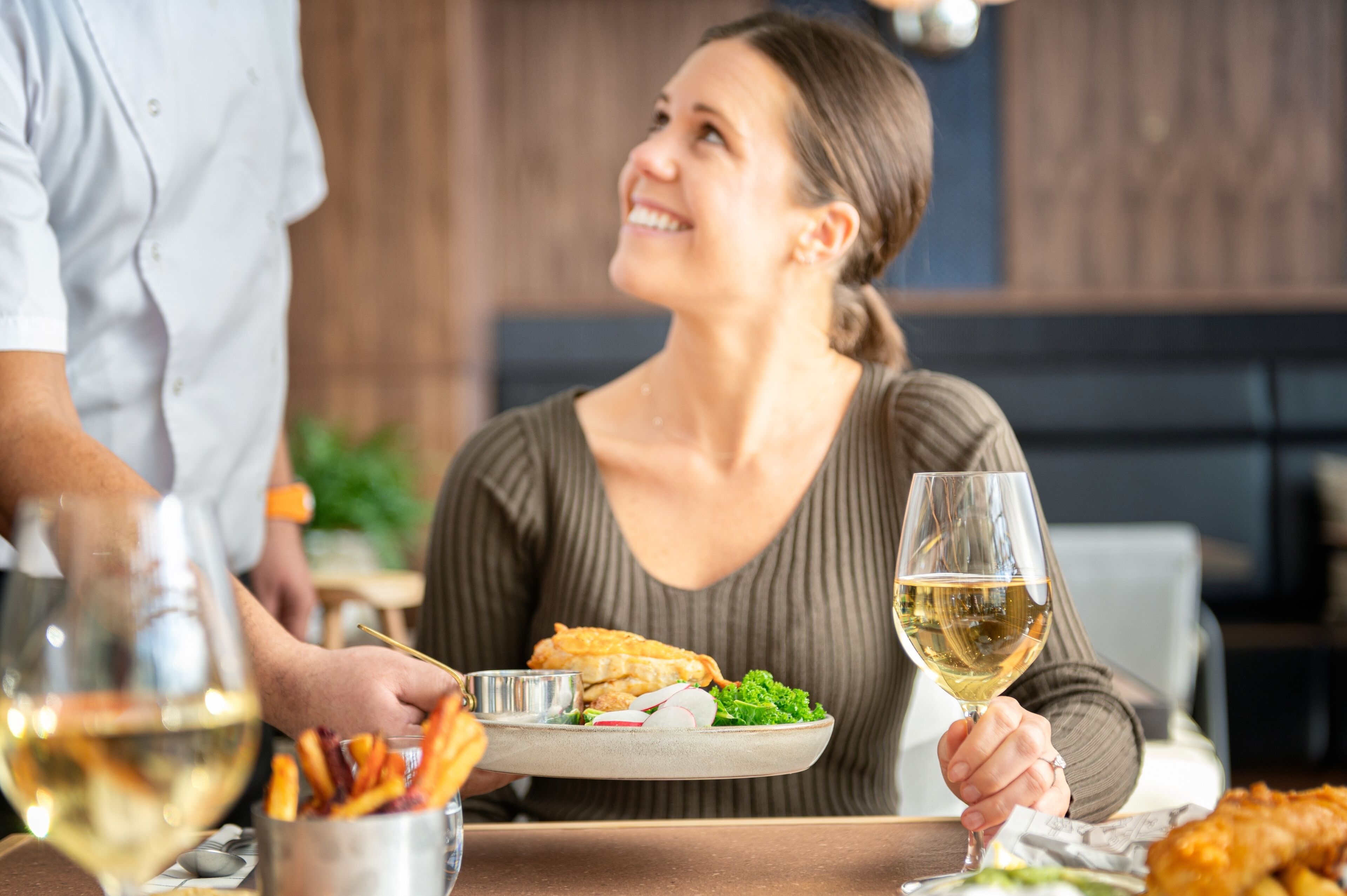 A dining table with a plate of food, a glass of white wine, and a person holding a fork.