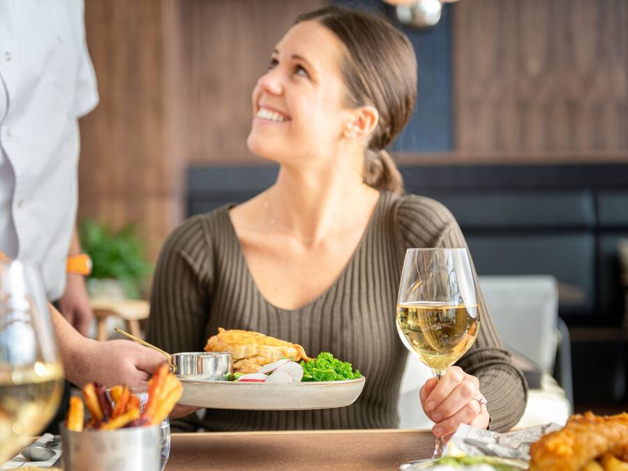 A dining table with a plate of food, a glass of white wine, and a person holding a fork.