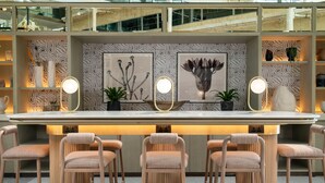 A modern bar area with a long counter, multiple stools, and wall-mounted lighting. - Hilton London Heathrow Airport Hotel (Hounslow)