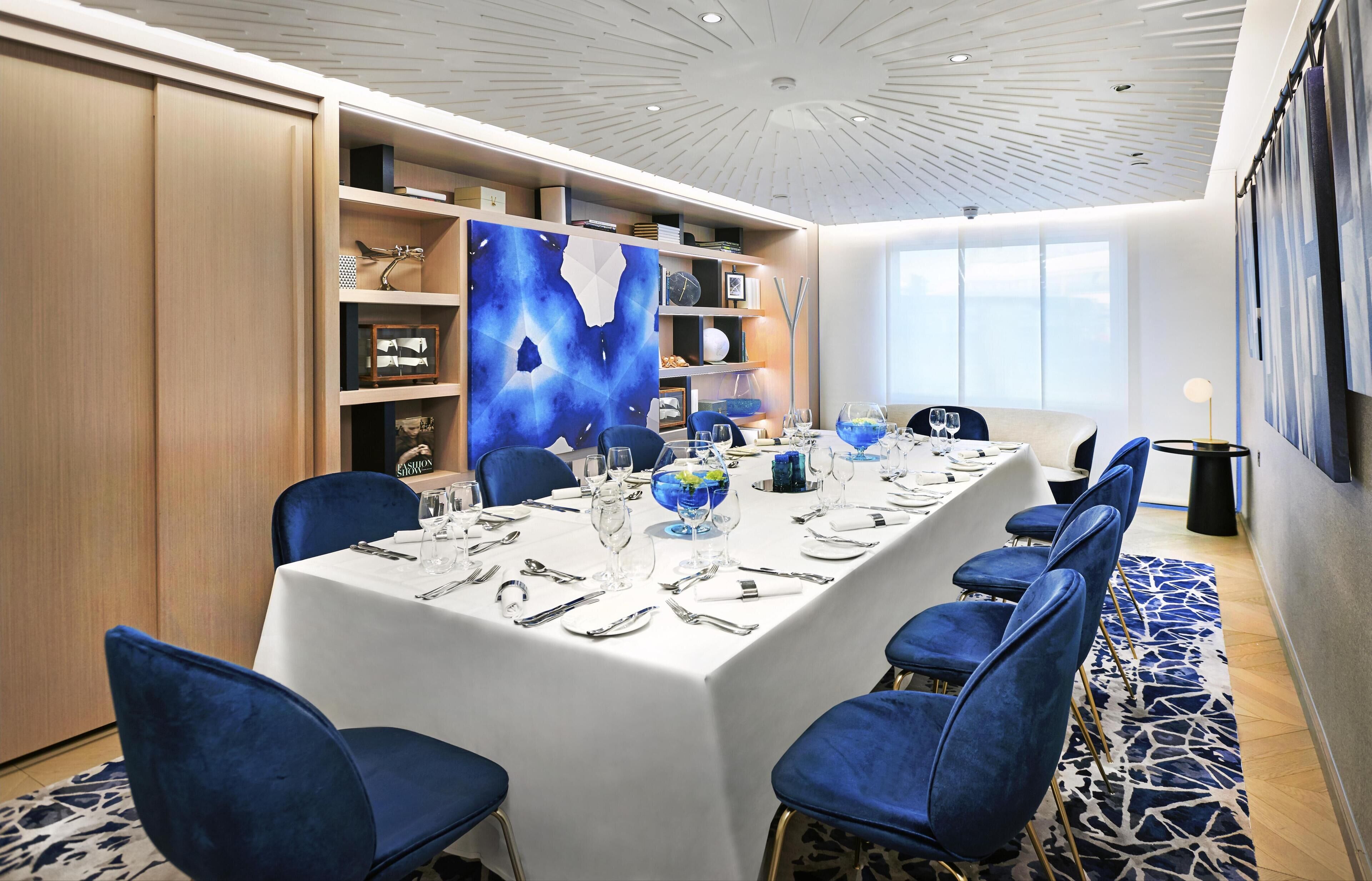 Un coin repas avec une longue table dressée pour six personnes, des chaises bleues et un grand tableau abstrait accroché au mur.
