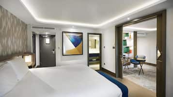 A hotel room with a large bed, a desk with a chair, a painting on the wall, and a door leading to a bathroom.