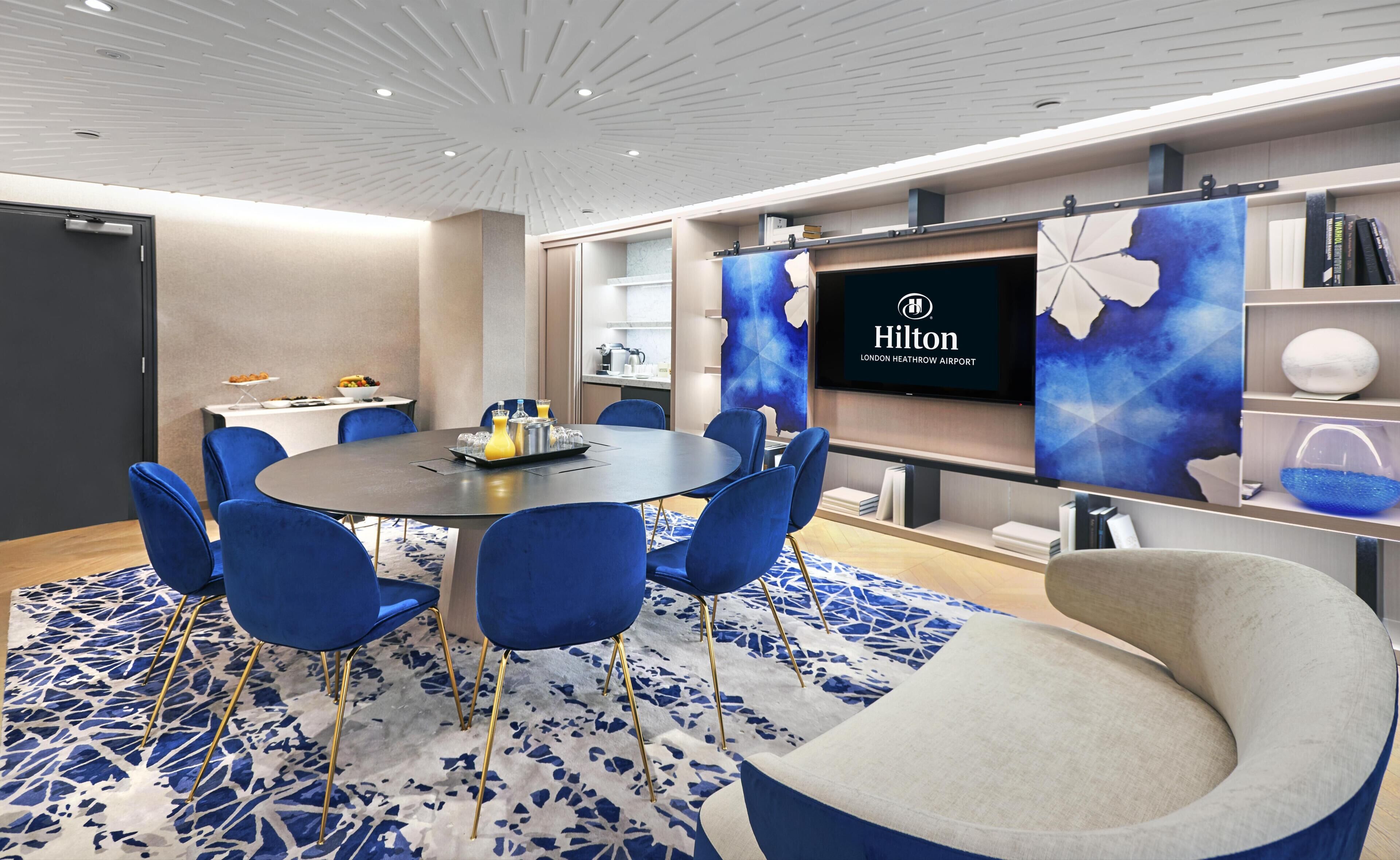 A modern conference room with a round table, blue chairs, a large screen displaying the Hilton logo, and a bookshelf with books and decorative items.
