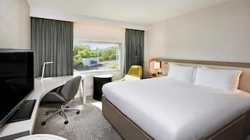 A hotel room with a bed, a desk with a chair, a TV, a window with curtains, and a nightstand with a lamp.