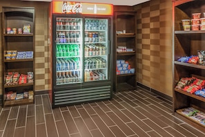 Máquina expendedora llena de bebidas y aperitivos, rodeada de estantes con productos envasados.