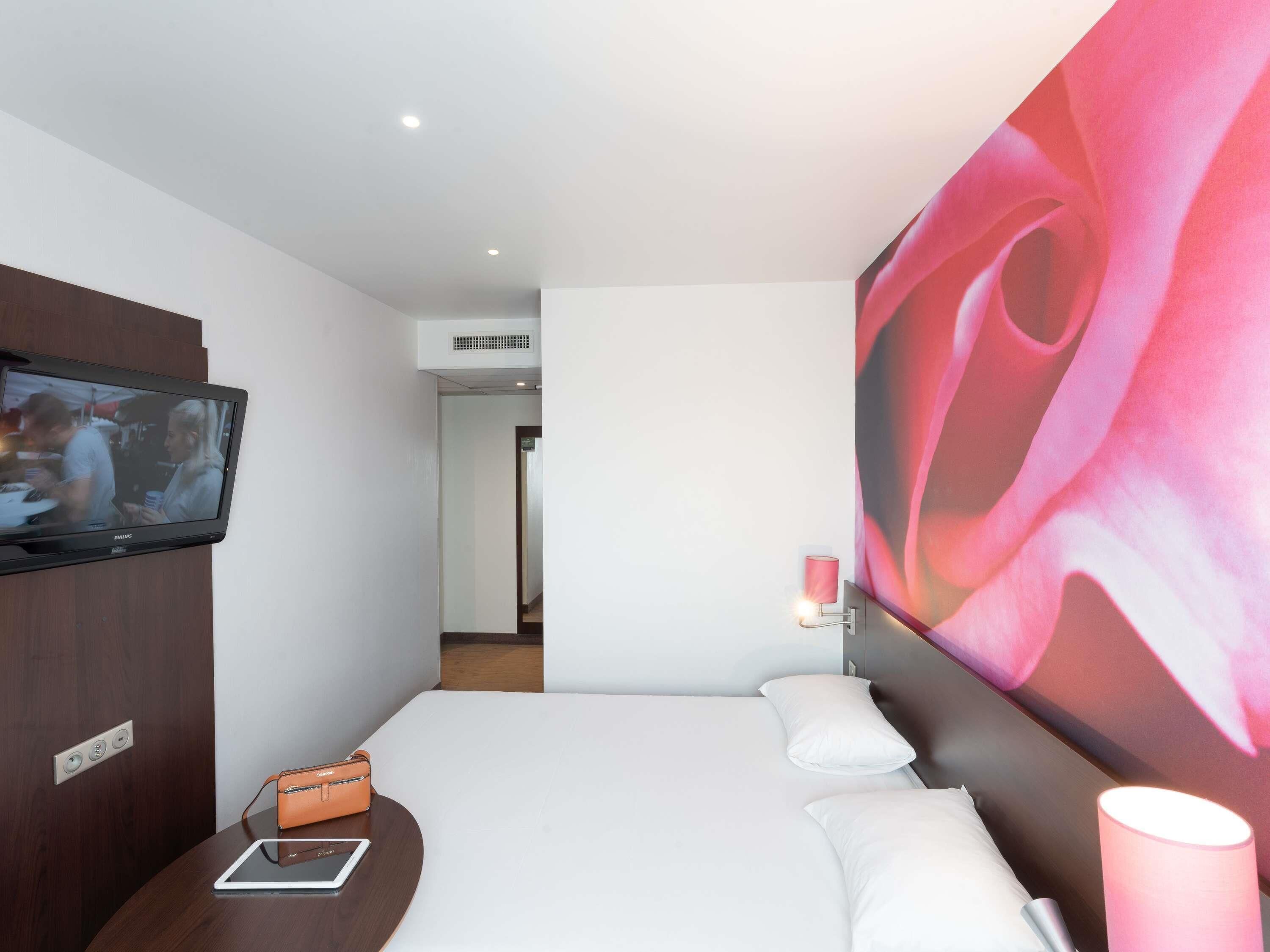 A hotel room with a bed, a TV mounted on the wall, a bedside table with a tablet, a small orange bag, and a large abstract painting on the wall.