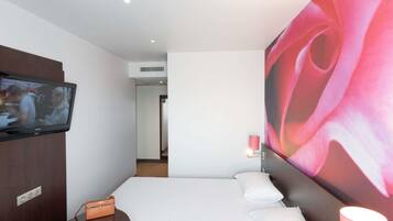 A hotel room with a bed, a TV mounted on the wall, a bedside table with a tablet, a small orange bag, and a large abstract painting on the wall.
