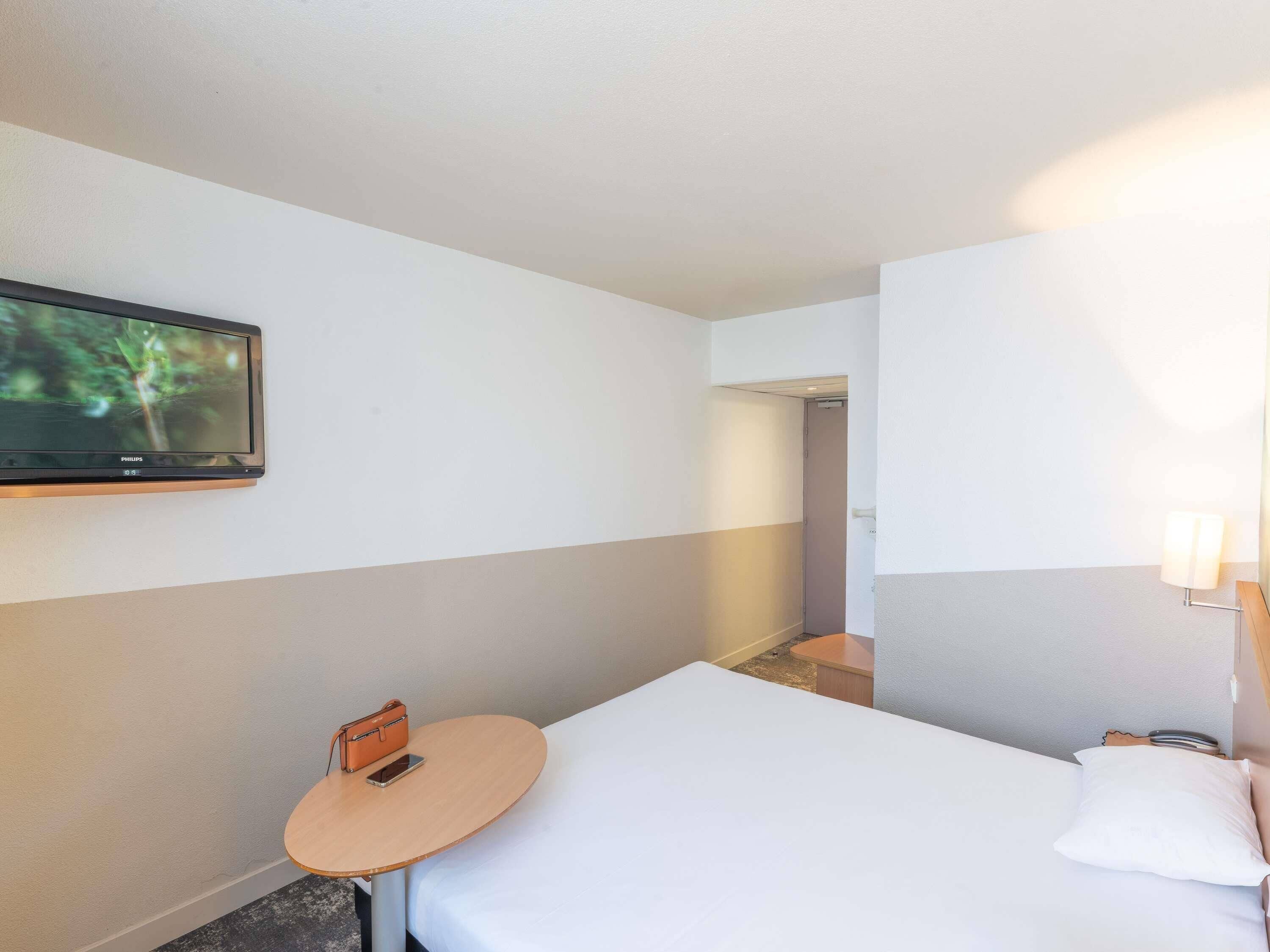 Une chambre d’hôtel avec un lit, une petite table avec un téléphone, un téléviseur fixé au mur et un miroir avec un éclairage intégré.