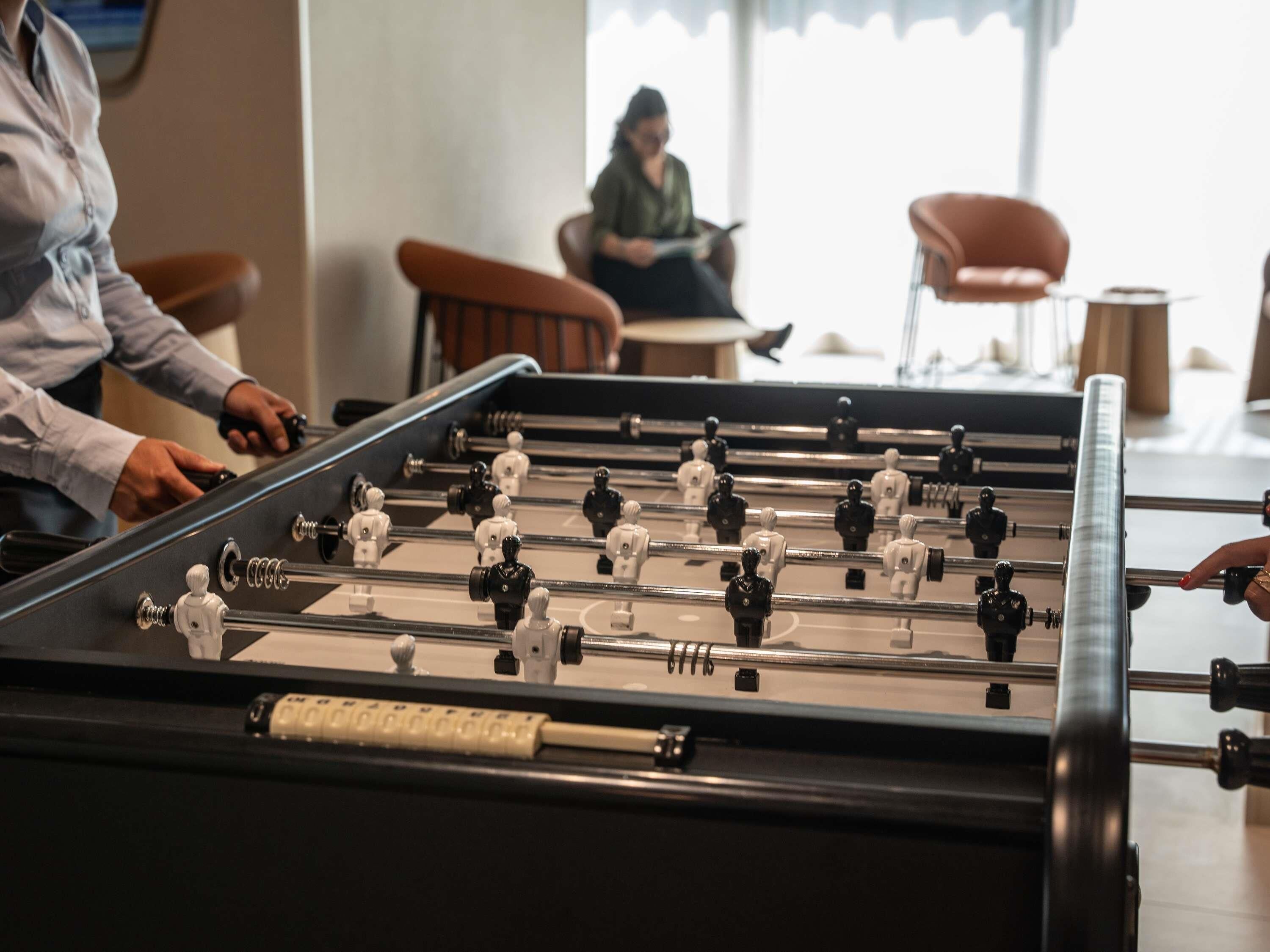 Une table de baby-foot dans une pièce avec des chaises, et une personne assise à l’arrière-plan.