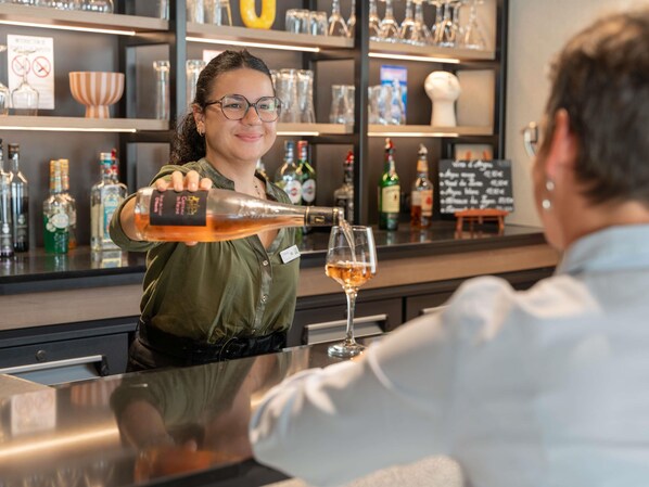 Een bartender schenkt een drankje in een glas aan een bar, met flessen en glazen achter haar.