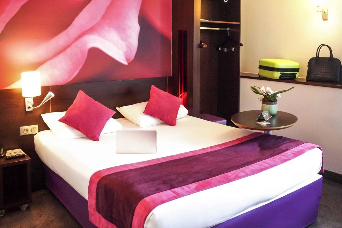 A hotel room with a bed, a nightstand, a desk, a wardrobe, and a small table with a plant.