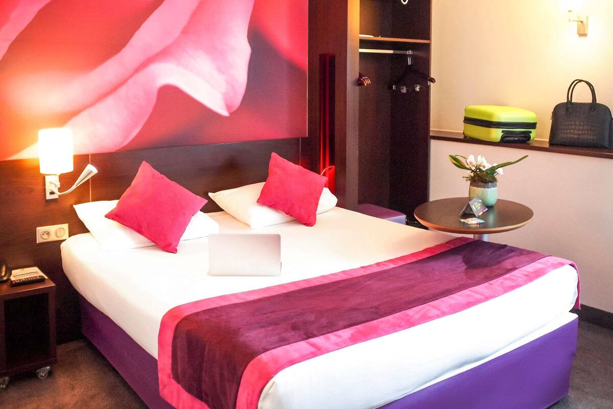 Une chambre d’hôtel comprenant un lit, une table de chevet avec une lampe, une petite table avec une plante et une armoire.