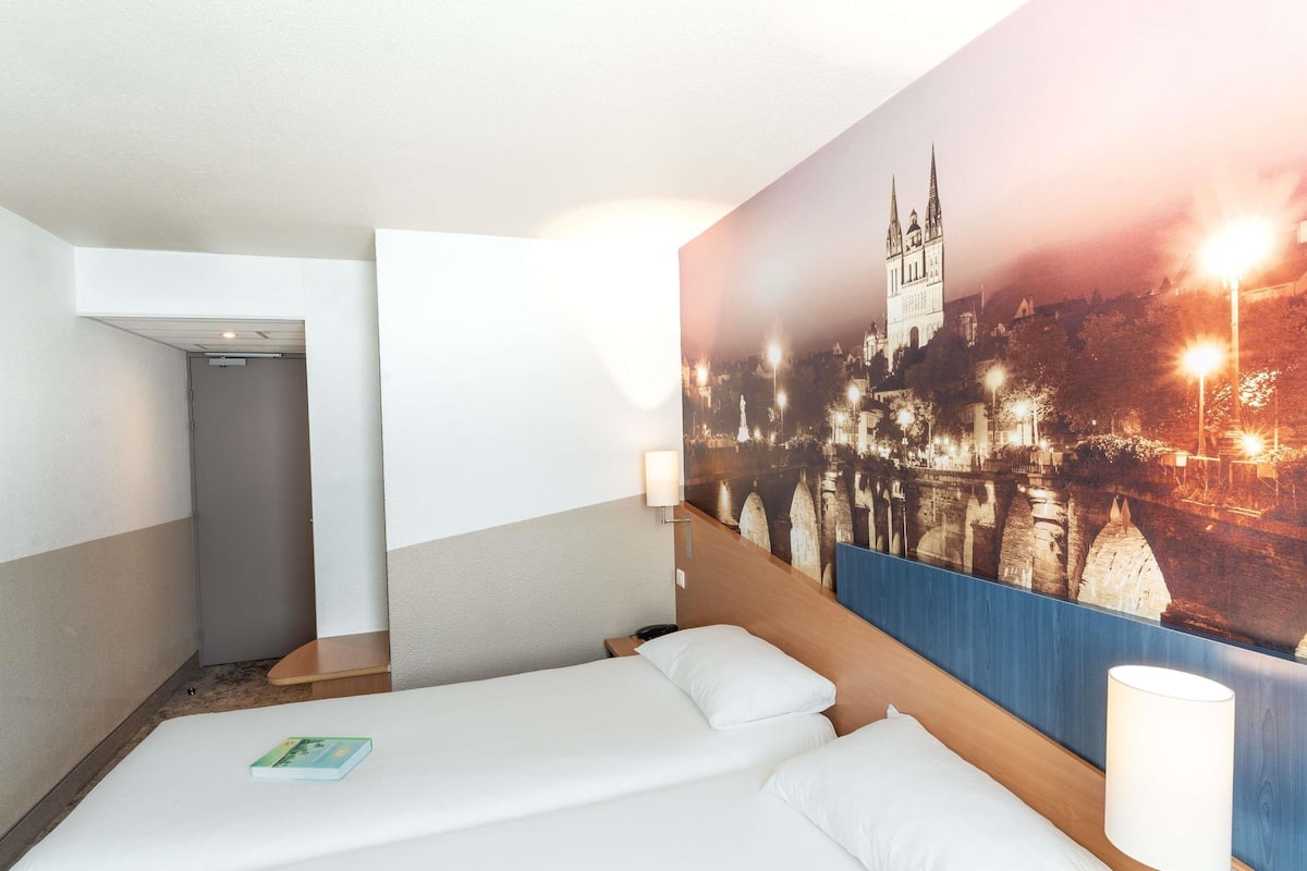 Une chambre d’hôtel avec deux lits, une table de chevet, une lampe fixée au mur et une grande fresque représentant un paysage urbain.