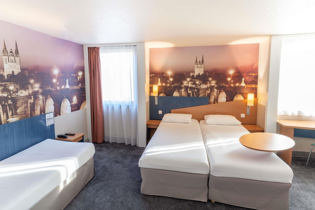 Une chambre d’hôtel avec deux lits, une petite table, une chaise, une fenêtre avec des rideaux et une fresque représentant un paysage urbain sur le mur.